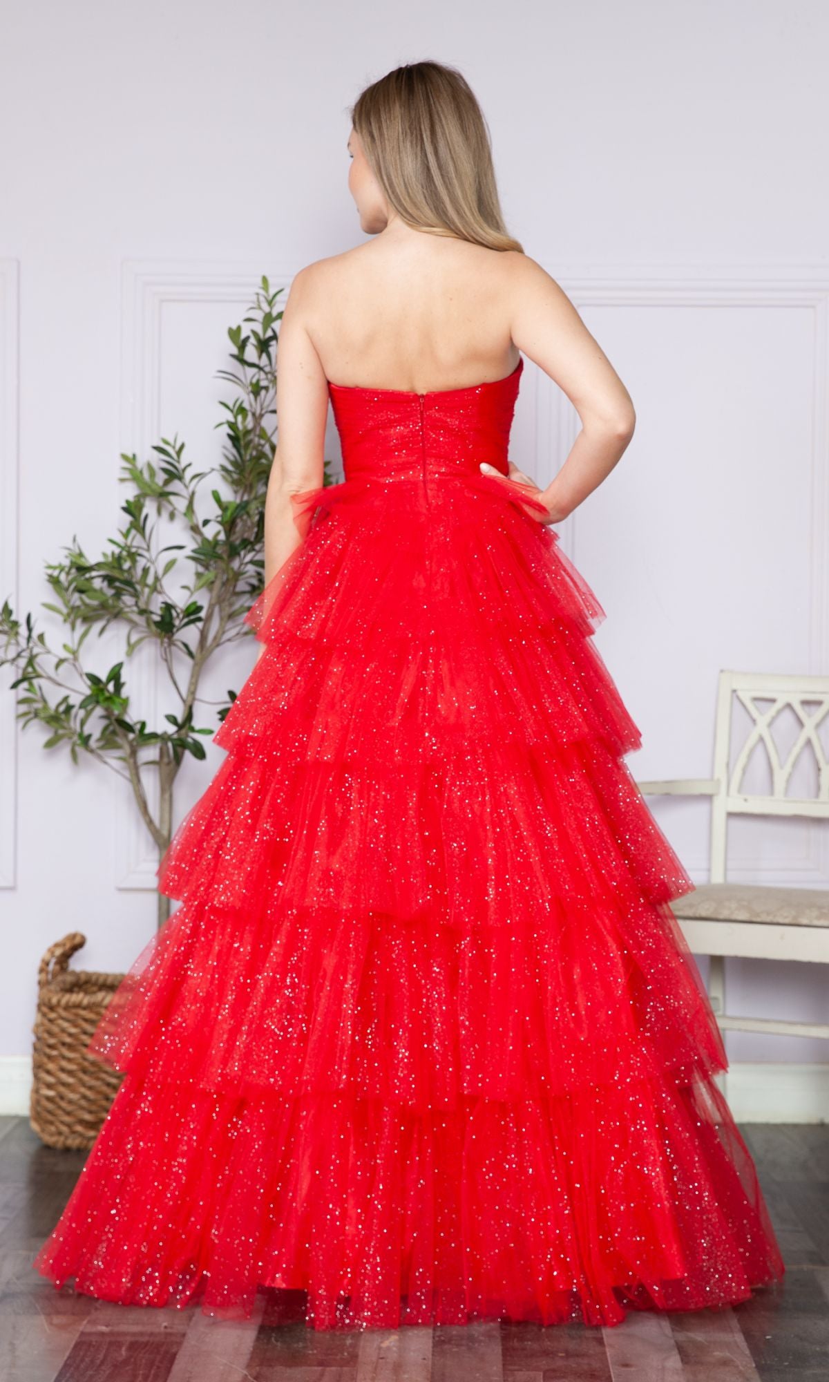 A woman with long blonde hair models the Strapless Long Tiered Glitter Prom Ball Gown 9386, standing with her back to the camera in a light-walled room with wooden floors.
